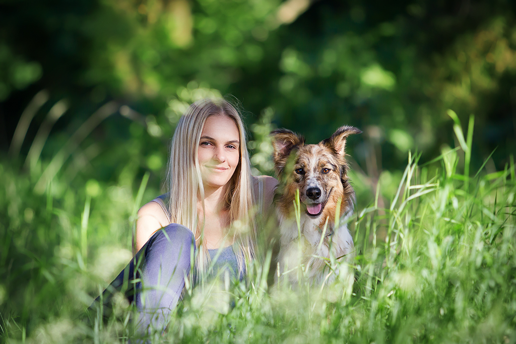 Ein blondes Mädchen sitzt mit ihrem Australian Shepherd im hohen Gras und schaut in die Kamera.